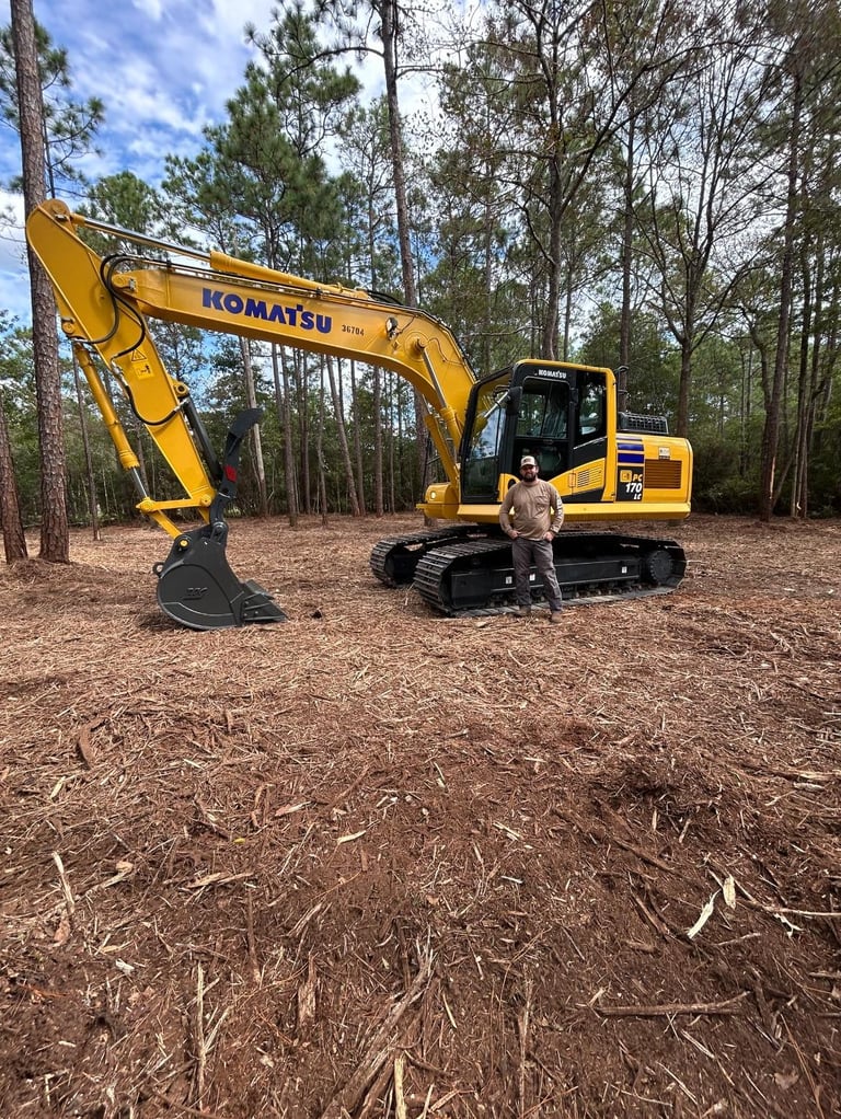 Person standing next to a yellow Komatsu excavator on a forest clearing with tall pine trees in the background