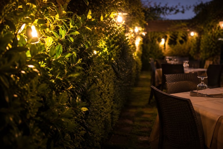 Patio esterno notturno con siepi illuminate e tavoli