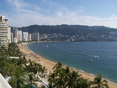 Aerial view of a crowded beach with high-rise hotels, palm trees, and turquoise water along a curved coastline with hills in the background