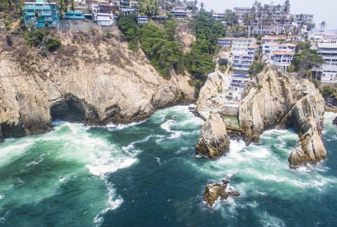 Aerial view of rocky coastal cliffs with turquoise ocean waves and residential buildings perched on the hillside above