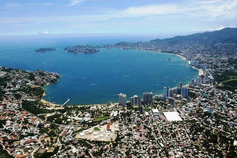 Aerial view of a coastal Mediterranean town with turquoise bay, sandy beaches, high-rise buildings, and forested hills