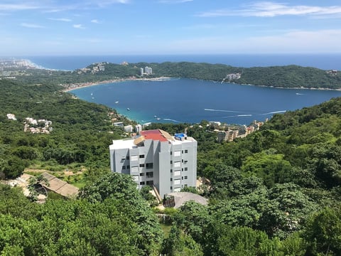 Modern white apartment building with red roof perched on hillside overlooking a blue bay surrounded by green forest and coastal landscape.