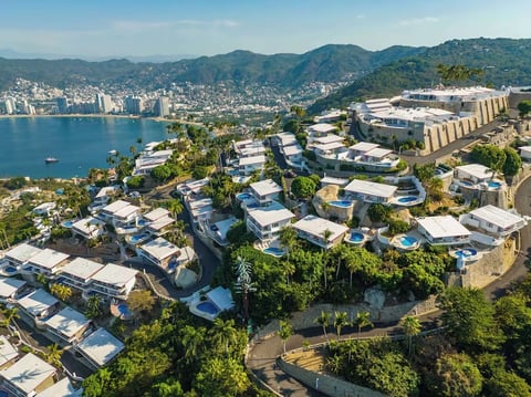 Aerial view of modern residential buildings on a hillside overlooking a blue coastal bay with mountains in the background