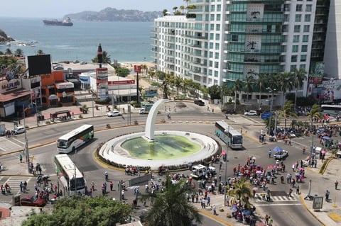 Aerial view of a coastal plaza with a circular white structure containing green water, surrounded by crowds, buildings, and ocean in the background