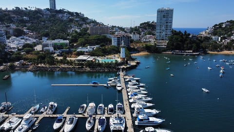 Aerial view of a coastal marina with white boats docked, surrounded by waterfront residential buildings and tree-covered hills