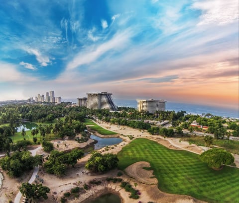 Aerial view of a coastal golf course with manicured greens, sandy bunkers, ocean, and high-rise buildings at sunset