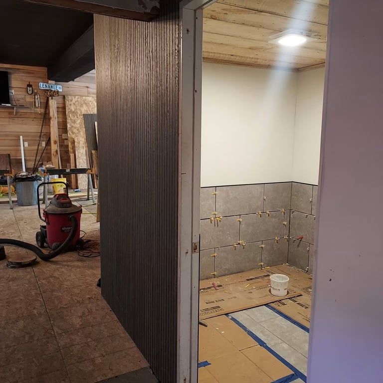 Construction site with an open-frame shower stall featuring corrugated metal walls and tiled interior under construction.