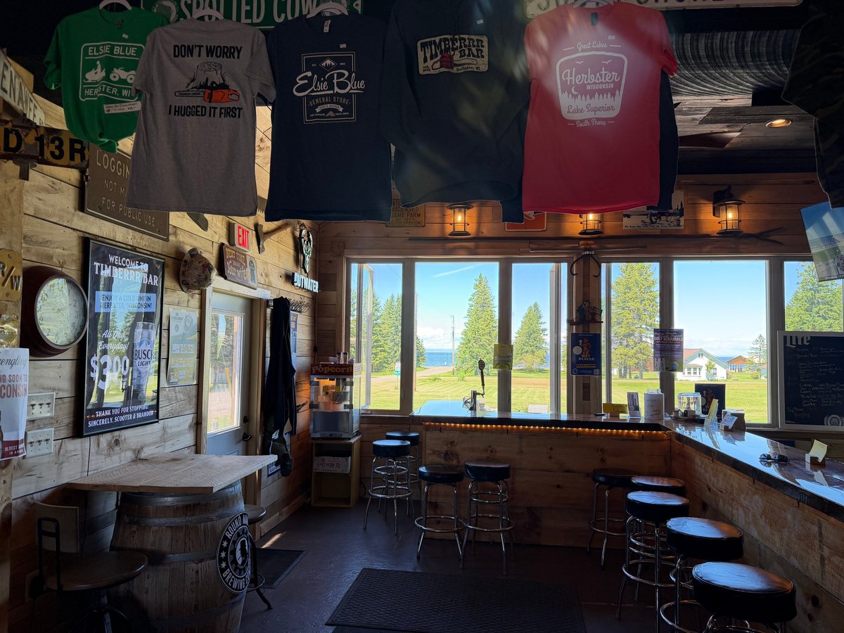 Bar interior with row of branded t-shirts hanging above the bar, stools lined up, and big windows looking out across the grass to Lake Superior