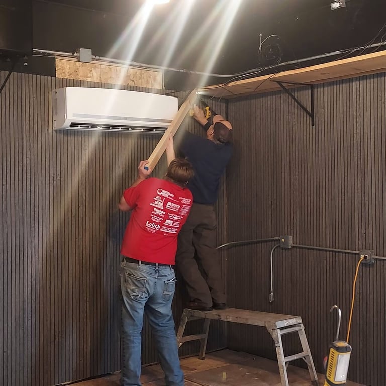 Two people installing a white air conditioning unit on a corrugated metal wall inside an industrial space.