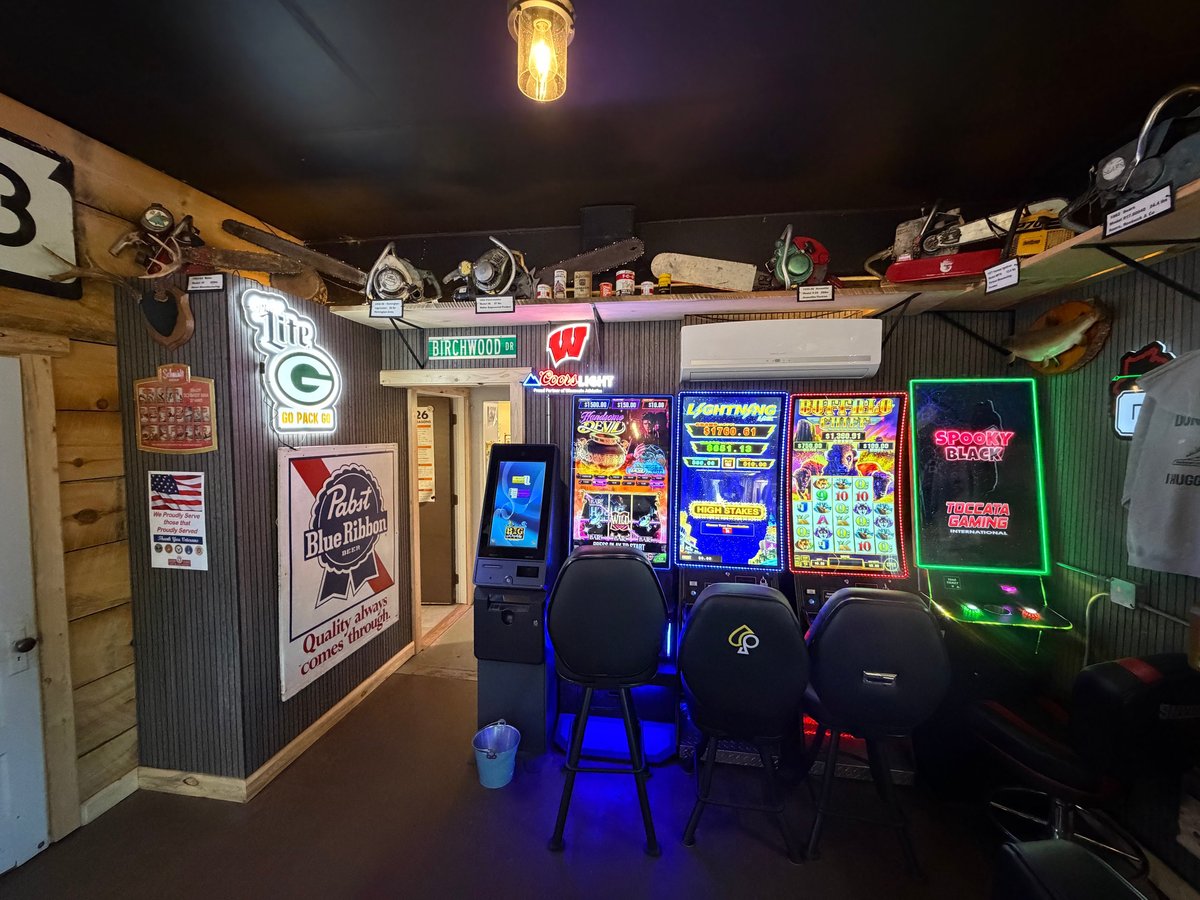 Game room corner with Pabst Blue Ribbon and Lite signs, three glowing slot machines, an ATM kiosk, and chainsaws displayed on high shelves