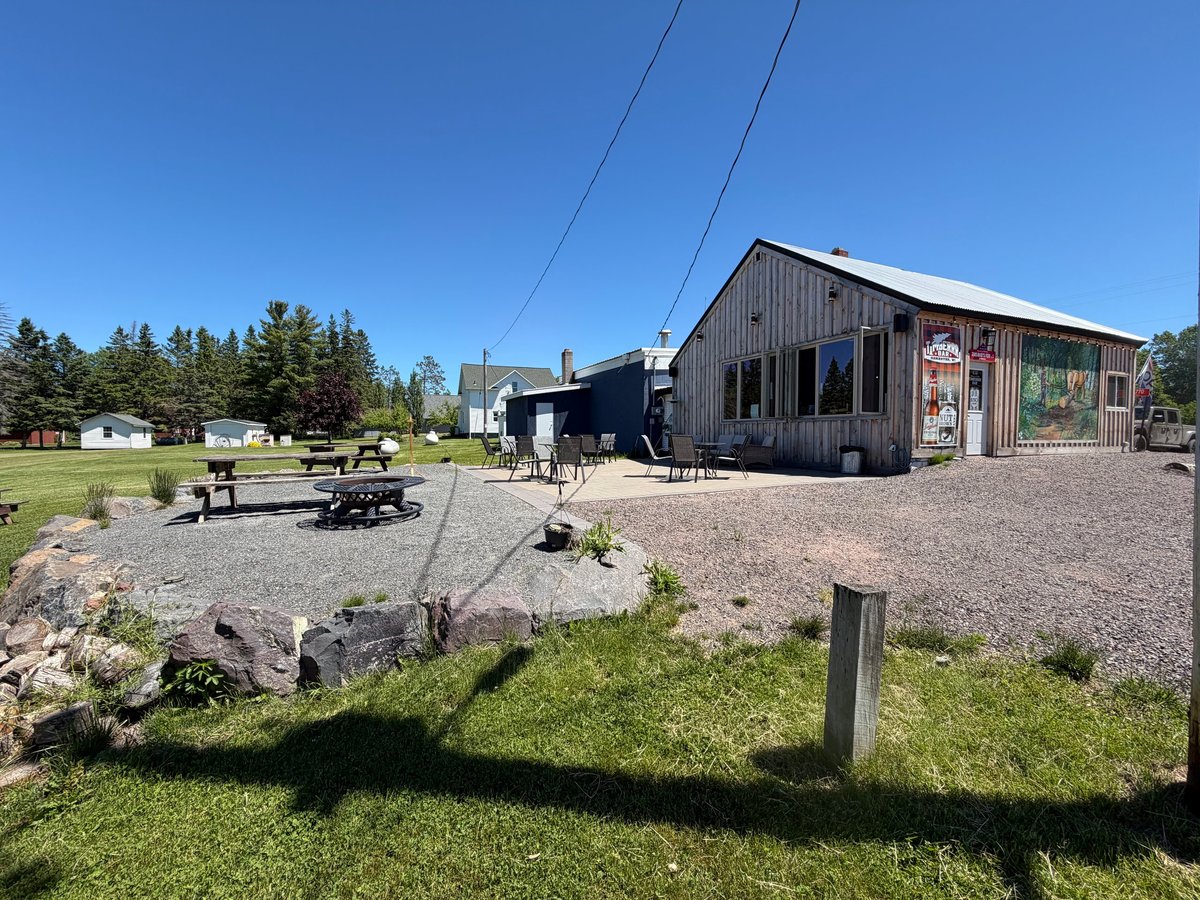 Rustic timber building with gravel lot, outdoor picnic area, and surrounding evergreen trees under bright blue Northwoods sky