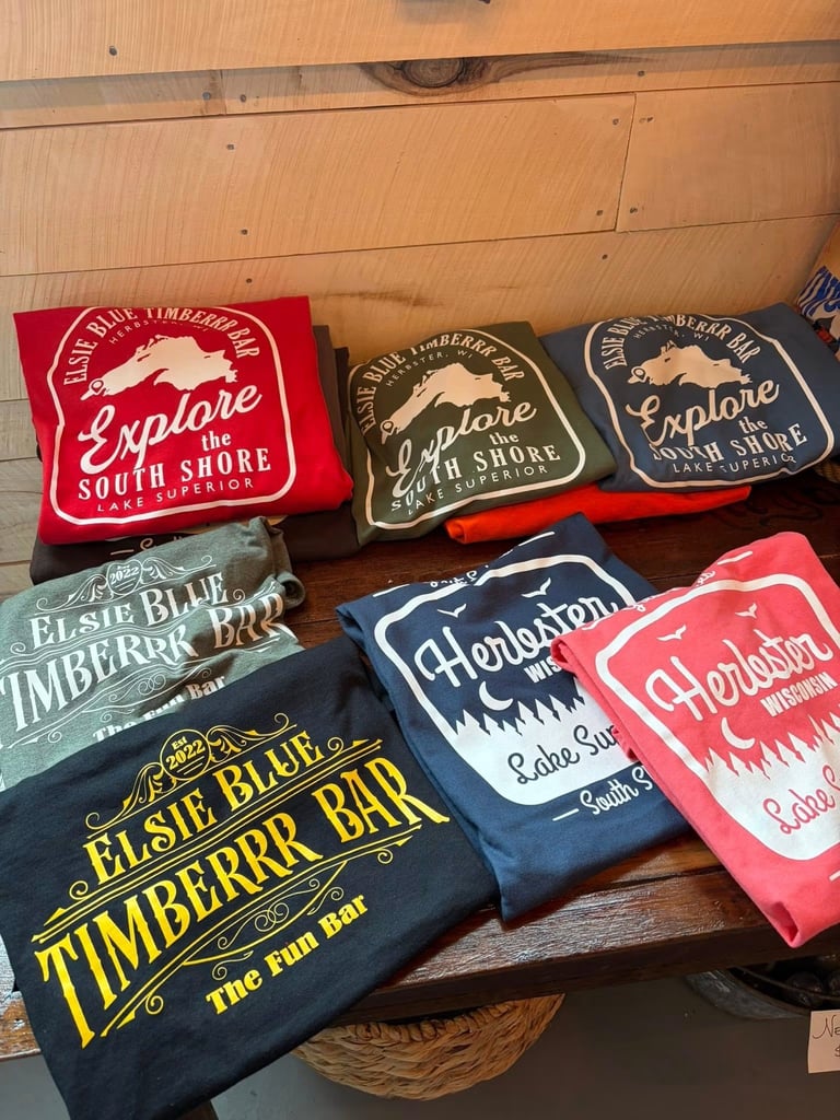 Collection of vintage-style travel souvenirs and apparel featuring Elsie Blue, Timberrr Bar, and Cheroladon South Shore designs displayed on wooden surface