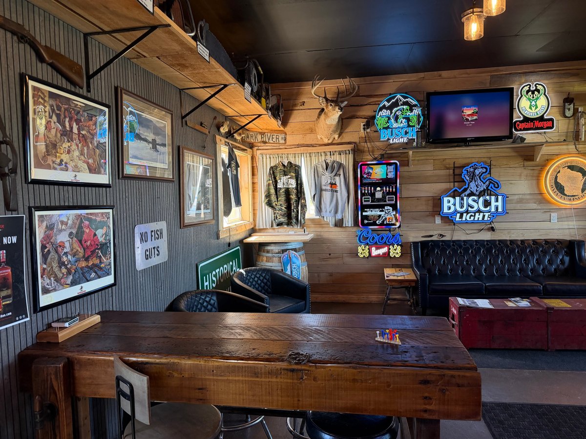 Rustic bar lounge with wood-paneled walls, neon Busch Light sign, framed wildlife art, deer mount, and dark leather sofa seating