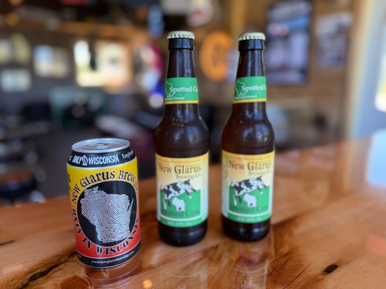 New Glarus Brewing Spotted Cow bottles and "Made in Wisconsin" can on a wooden bar counter, showcasing Wisconsin beer