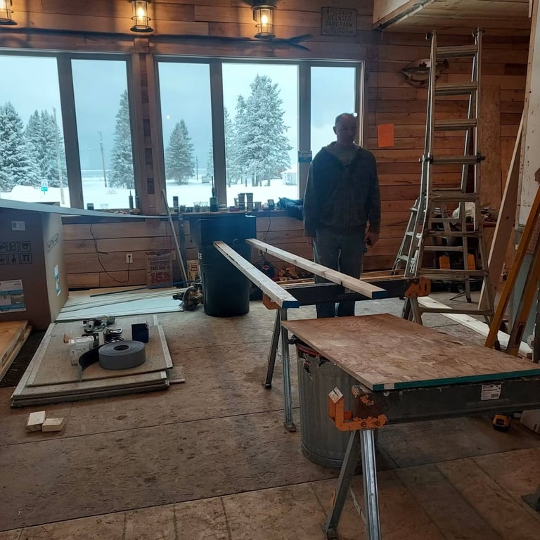 Construction worker in an unfinished cabin interior with tools, lumber, and snow-covered trees visible through large windows