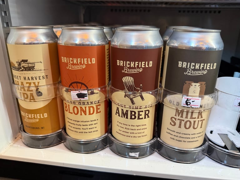 Four Brickfield Brewing beer cans from Grantsburg WI on a cooler shelf: Sunset Harvest Hazy IPA, Good Orange Blonde, Right Time Ritual Amber, and Old Milwaukee Filled Milk Stout