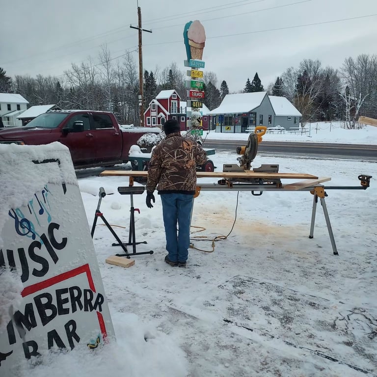 Person working at outdoor woodworking station in snowy rural setting with buildings and ice cream cone sign in background