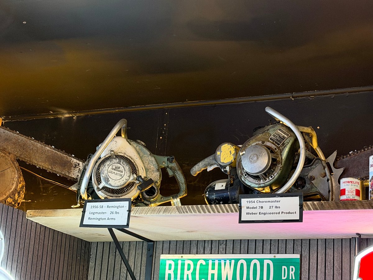 Two antique chainsaws displayed on a wood beam with informational placards — Remington Logmaster and Choremaster Model 3B at the Timberrr Bar