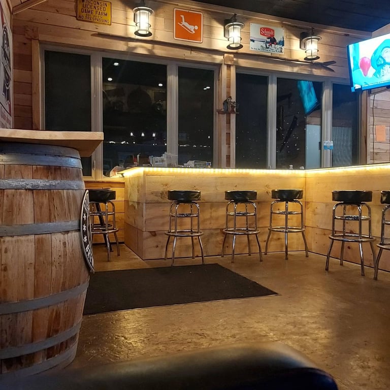 Interior of a rustic bar with wooden counter, bar stools, barrels, glowing signage, and large windows at night