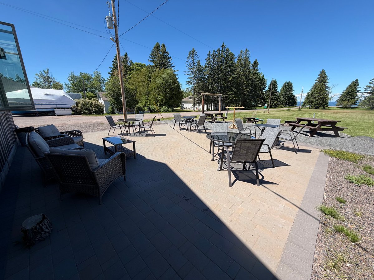 Outdoor patio at the Timberrr Bar with wicker chairs, bistro tables, and picnic tables, surrounded by tall pines with Lake Superior in the distance