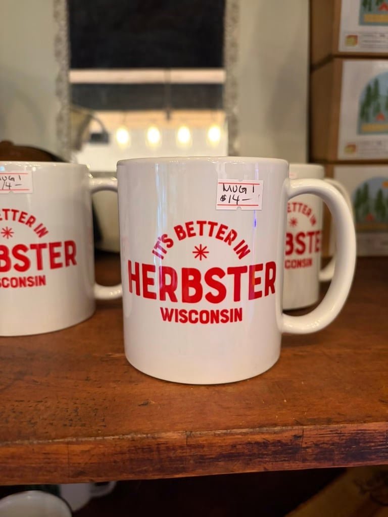 White ceramic mugs with red text reading Its Better in Herbster Wisconsin displayed on a wooden surface