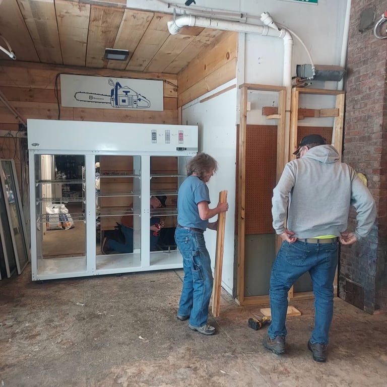 Two workers installing a large white cabinet or display case in an industrial workshop with exposed wood and brick.