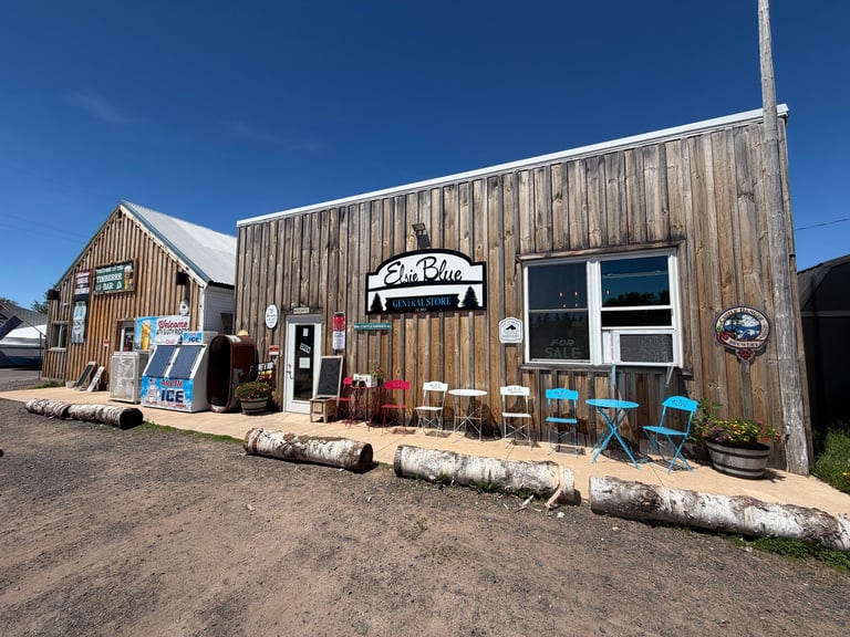 Rustic Elsie Blue General Store entrance with weathered wood siding, turquoise cafe chairs on the porch under clear blue sky