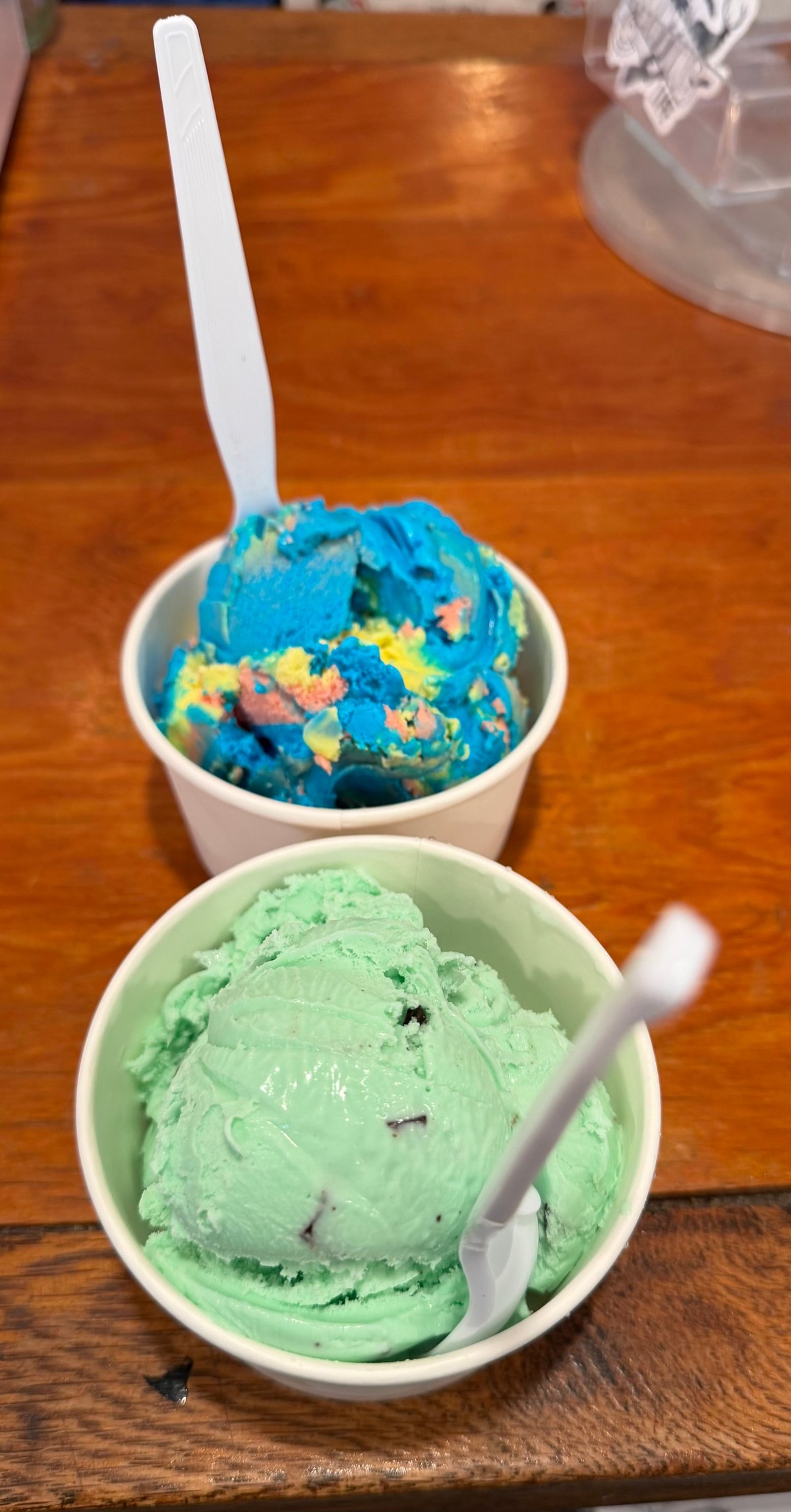 Two scoops of ice cream in white bowls on a wooden table — bright blue with colorful sprinkles, and pale green mint chip