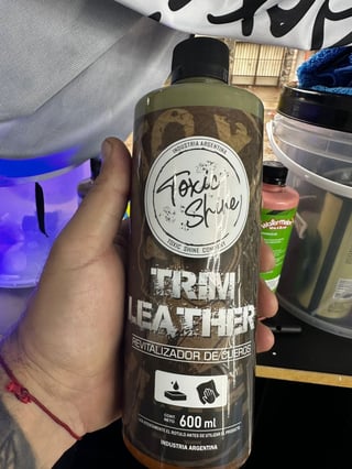 Hand holding a bottle of Toxic Shine Trim Leather cleaner with 600ml capacity, shown in an indoor setting with various products in the background