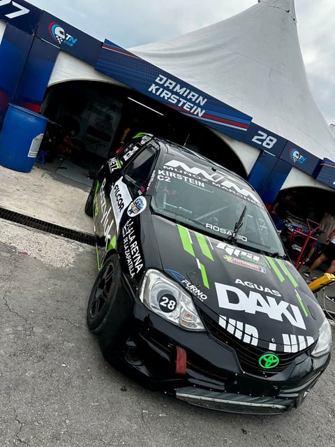 Black race car with green accents parked in front of blue team garage with number 28 visible
