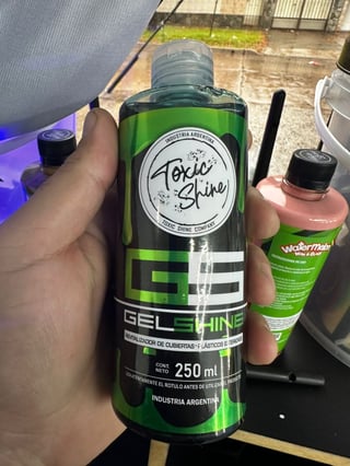 Hand holding a 250ml green Toxic Shine gel shine bottle with black label, with cleaning products nearby