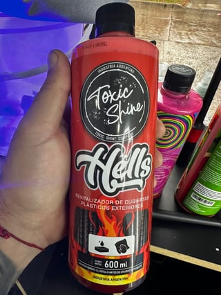 Hand holding a red 600ml bottle of Axe Shine Hell plastic restorer spray with colorful beauty products in background