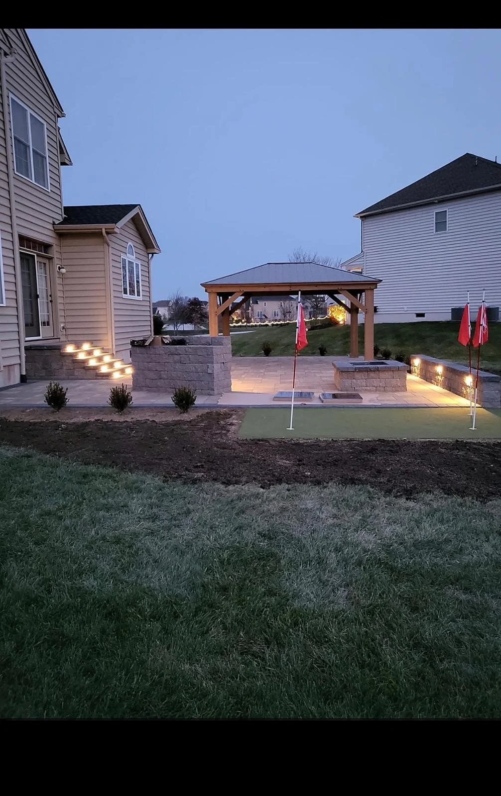 Backyard patio at dusk with illuminated stone seating area, wooden gazebo, and homes with manicured lawn