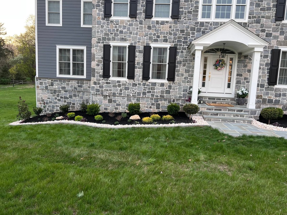 Two-story brick and stone house with white trim, black shutters, front entrance, landscaped garden beds, and manicured lawn