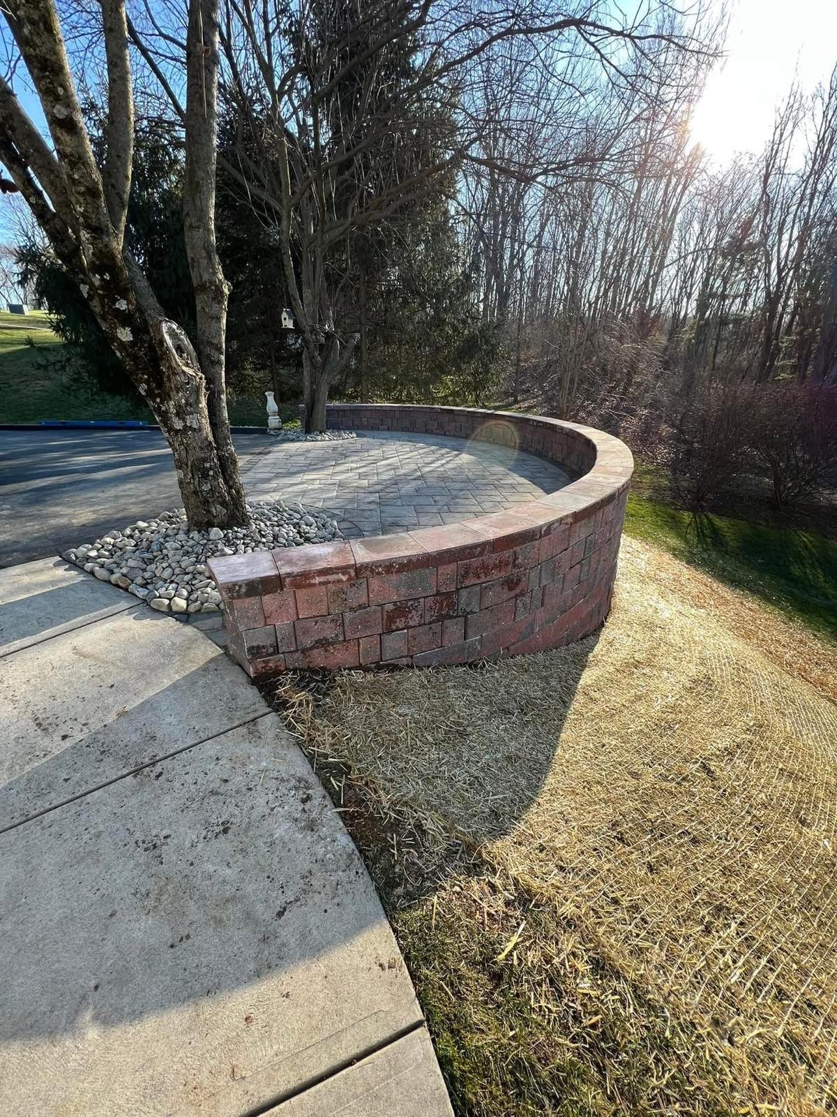 Curved red brick retaining wall with paved seating area next to bare trees in a landscaped yard