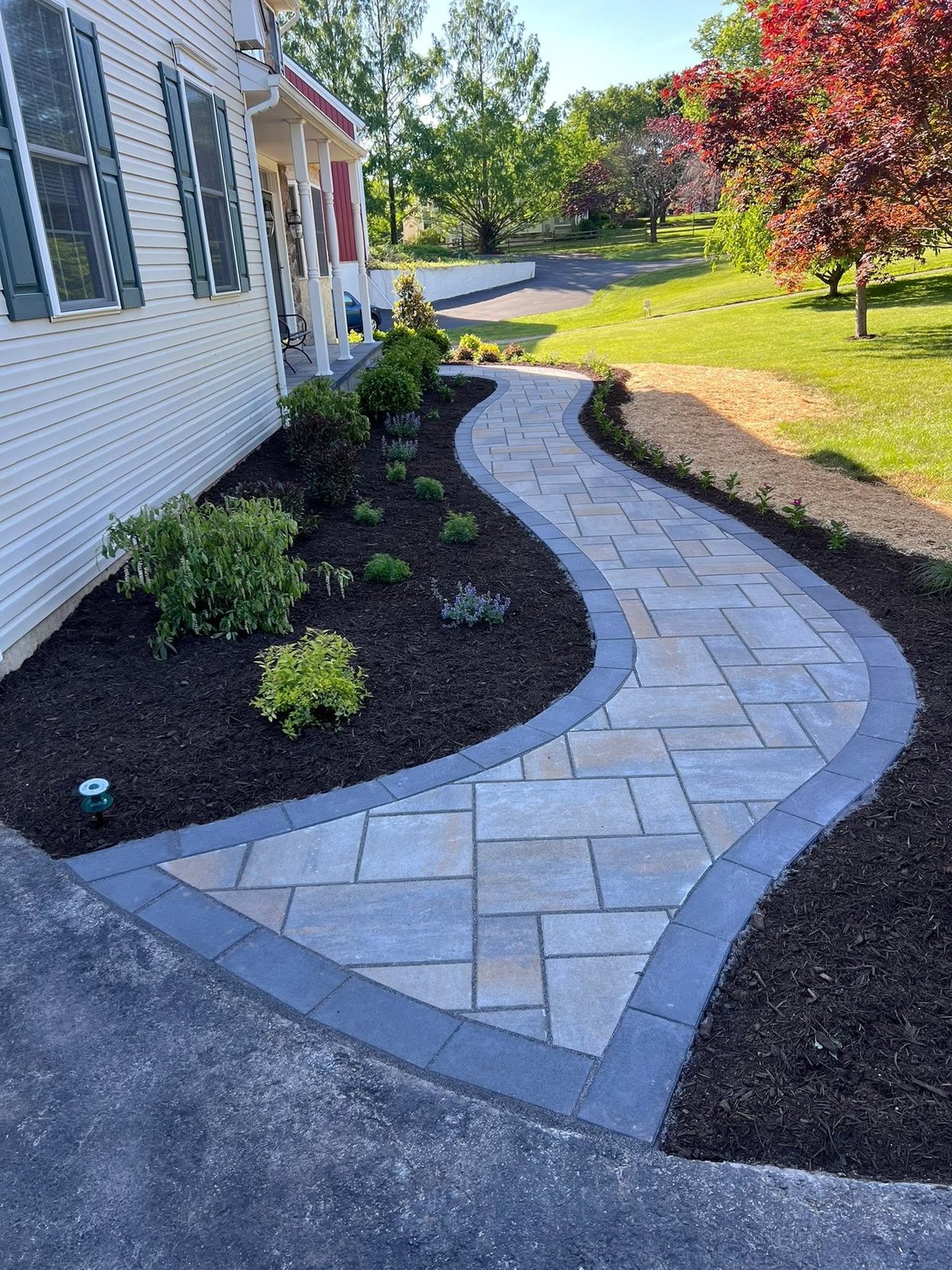 Curved paver walkway with landscaping next to white house and expansive lawn with mature trees