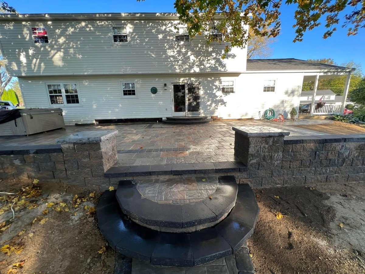 Backyard with new paver patio under construction, stone fire pit feature, and white house with garage in background