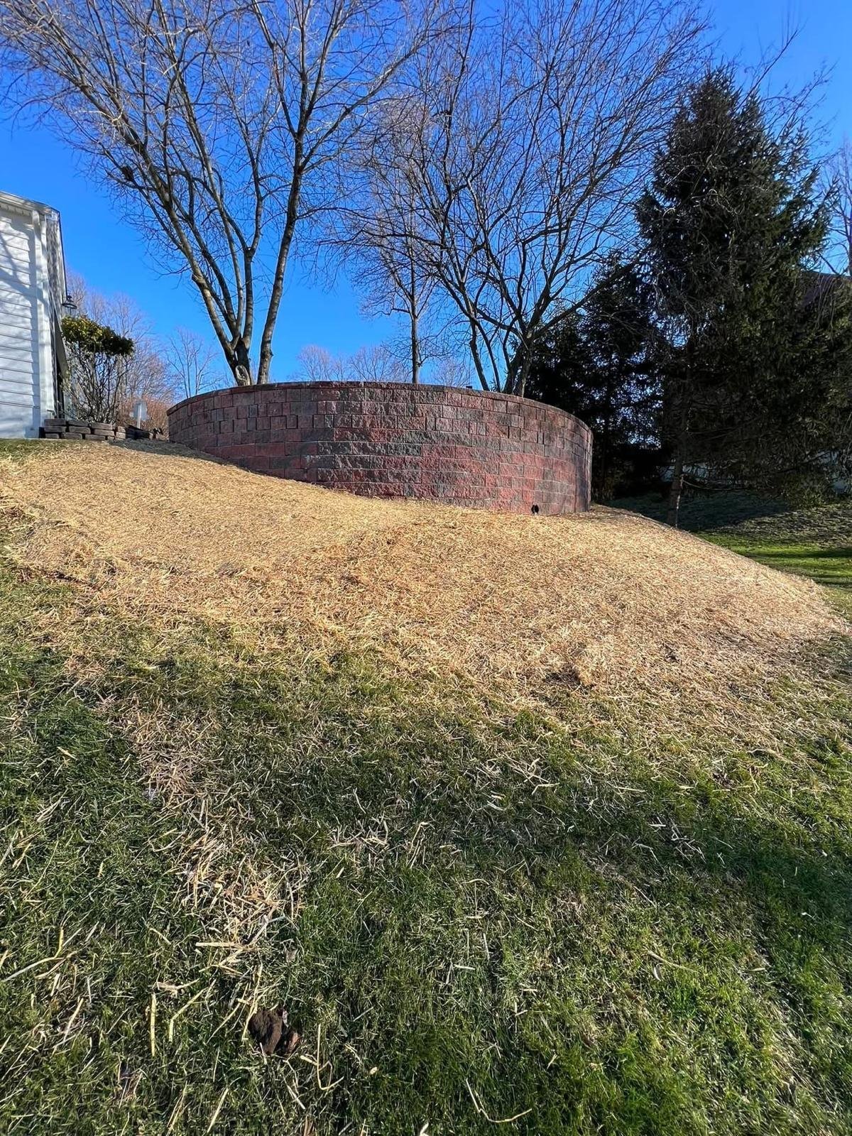 Brick storage silo on a grassy hillside with bare trees and a house visible on the left under clear blue sky