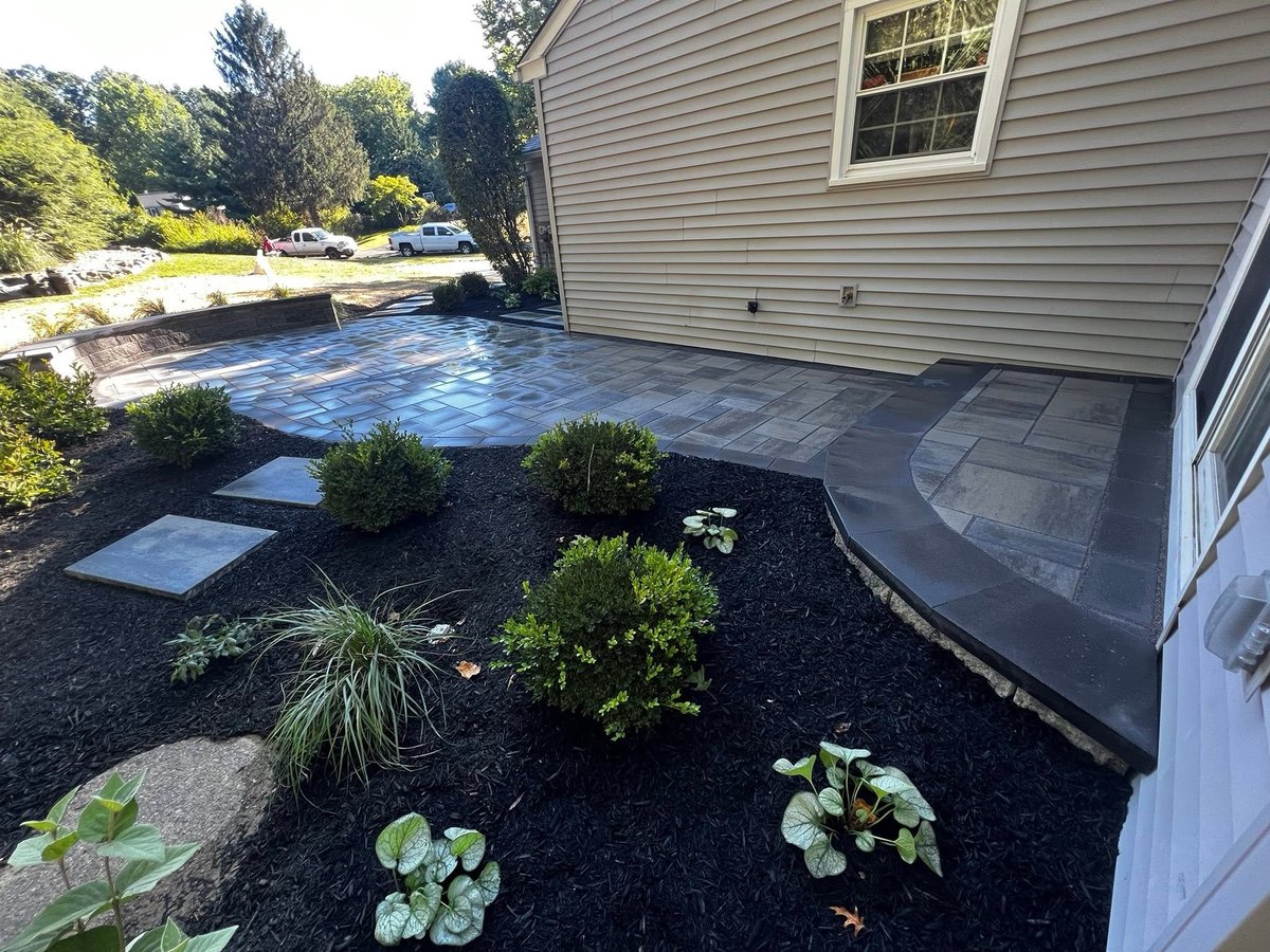 Backyard patio with blue-gray pavers, landscaped garden beds with shrubs and plants, gray siding house, and parked cars visible in background