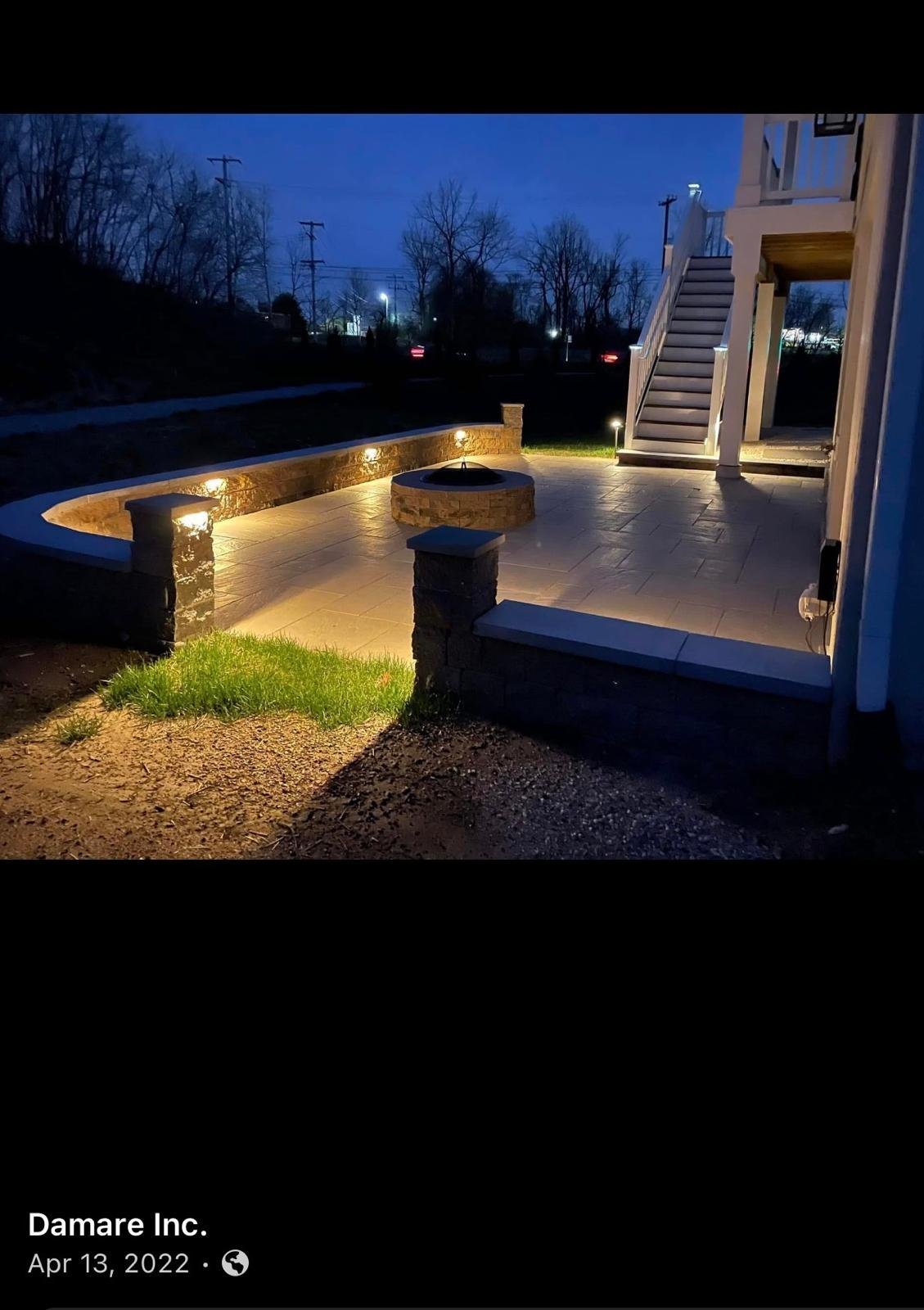 Illuminated outdoor patio and pool deck at dusk with curved seating, stone accents, and residential stairs alongside a modern home