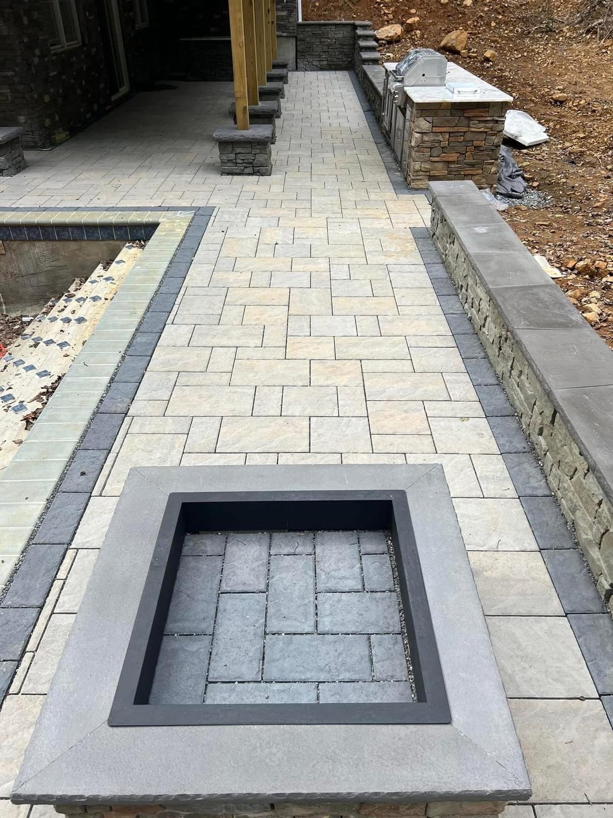 Paved backyard patio with geometric brick pattern, square fire pit insert, and stone walls during construction