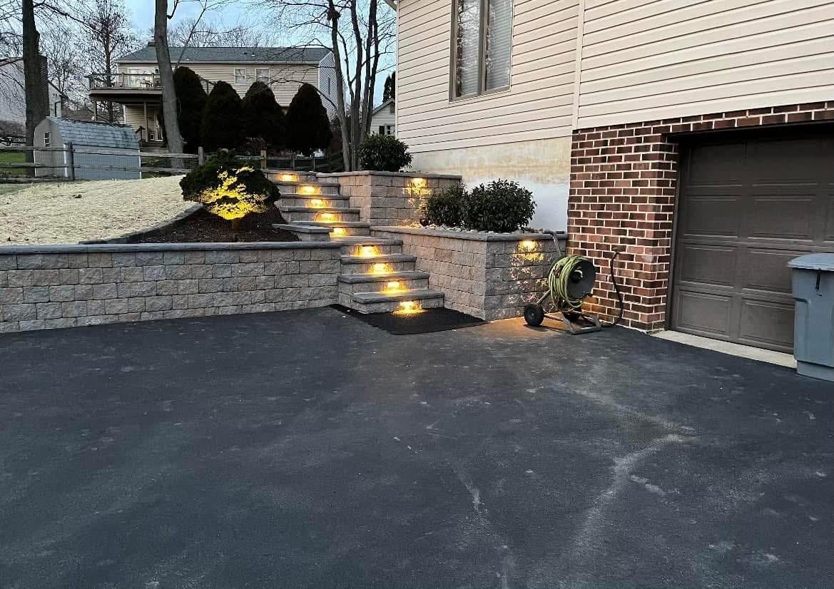 Residential driveway with illuminated stone retaining wall steps at dusk, brick home and garage visible