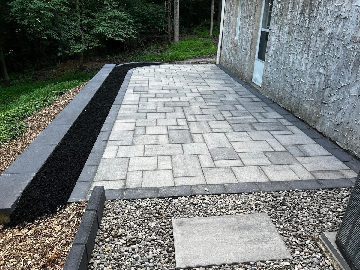 A newly installed paver patio with light gray pavers adjacent to a concrete building with black stone landscaping border