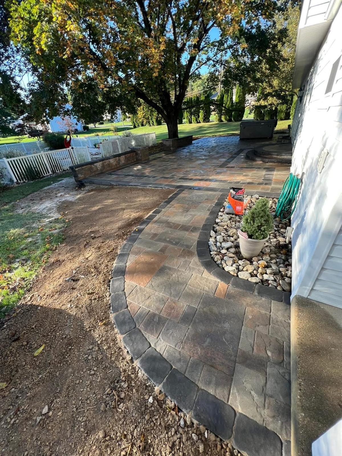 Curved brick patio next to white house with potted flowers, overlooking manicured lawn and water in background