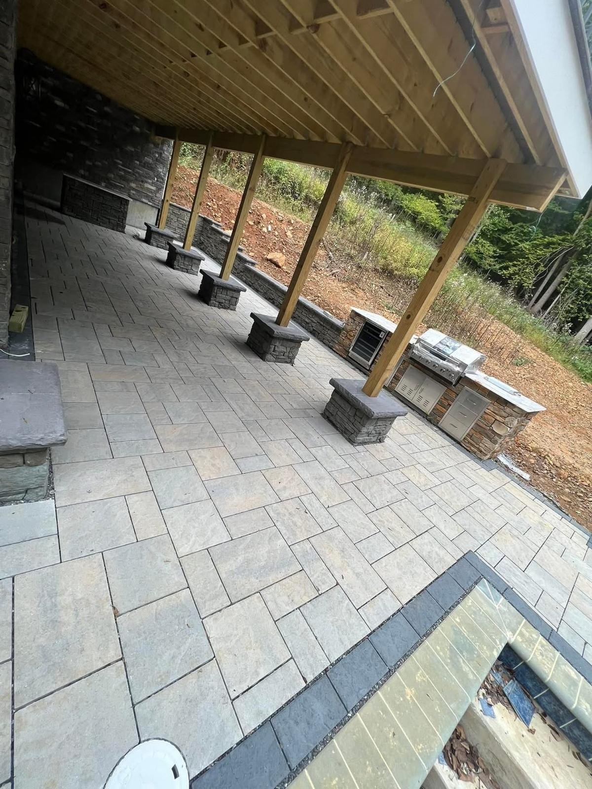 Covered outdoor patio with stone flooring, wooden beam structure, and metal fire pits under construction