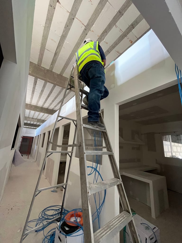 Técnico con chaleco y arnés sobre una escalera durante la instalación de infraestructura en una obra, con cableado y canalizaciones expuestas