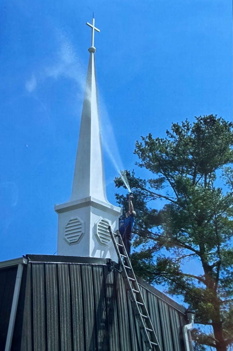 White church steeple with cross against blue sky, metal roof visible below with ladder leaning against it and tall tree nearby