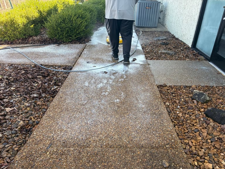 Person in light blue shirt pressure washing a wet concrete pathway between landscaped gravel areas near a building