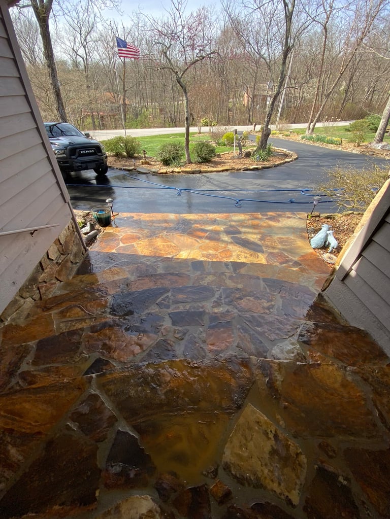 Wet flagstone walkway and asphalt driveway between two houses during a cleaning job — truck visible on road with American flag in background