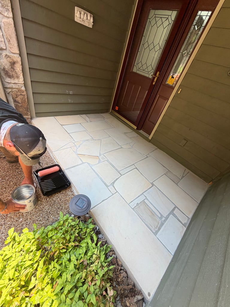 Worker laying light-colored tile on porch entrance with dark red door and green siding walls